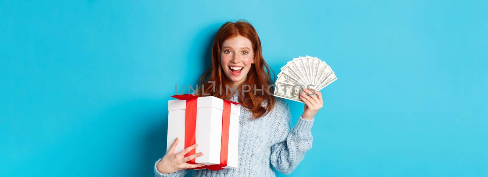 Christmas and shopping concept. Happy redhead woman holding money and big xmas present, showing dollars and gift, smiling pleased, standing over blue background.