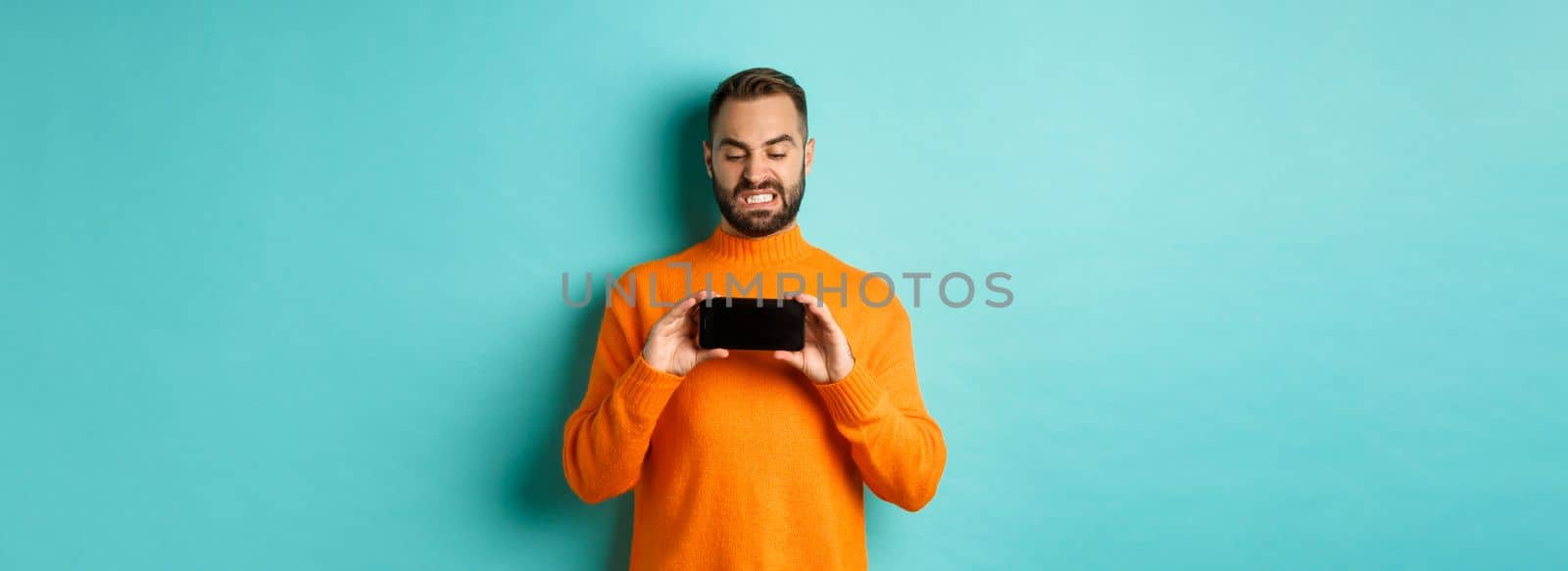 Image of man cringe at something on mobile screen, staring disgusted at display, showing smartphone, standing over light blue background by Benzoix