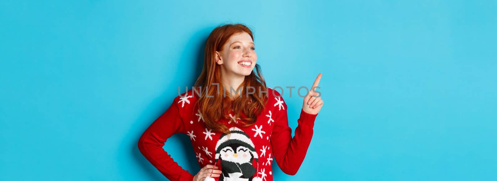 Winter holidays and Christmas Eve concept. Happy and cute redhead girl in xmas sweater, looking and pointing at upper left corner amazed, standing over blue background by Benzoix
