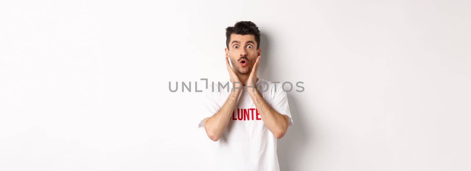 Shocked man in volunteer t-shirt gasping, staring at camera in awe, standing over white background.