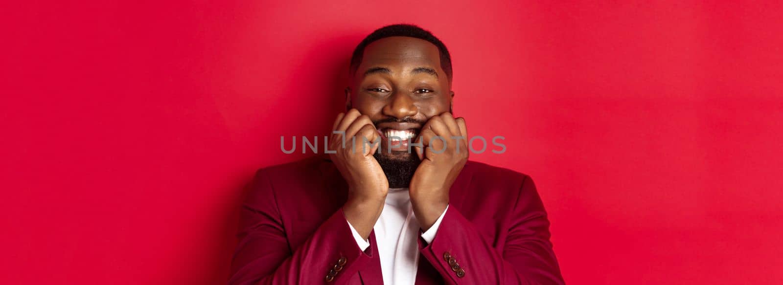 Close-up of silly african american man watching something adorable, touching cheeks and smiling with delight and happiness, standing over red background.