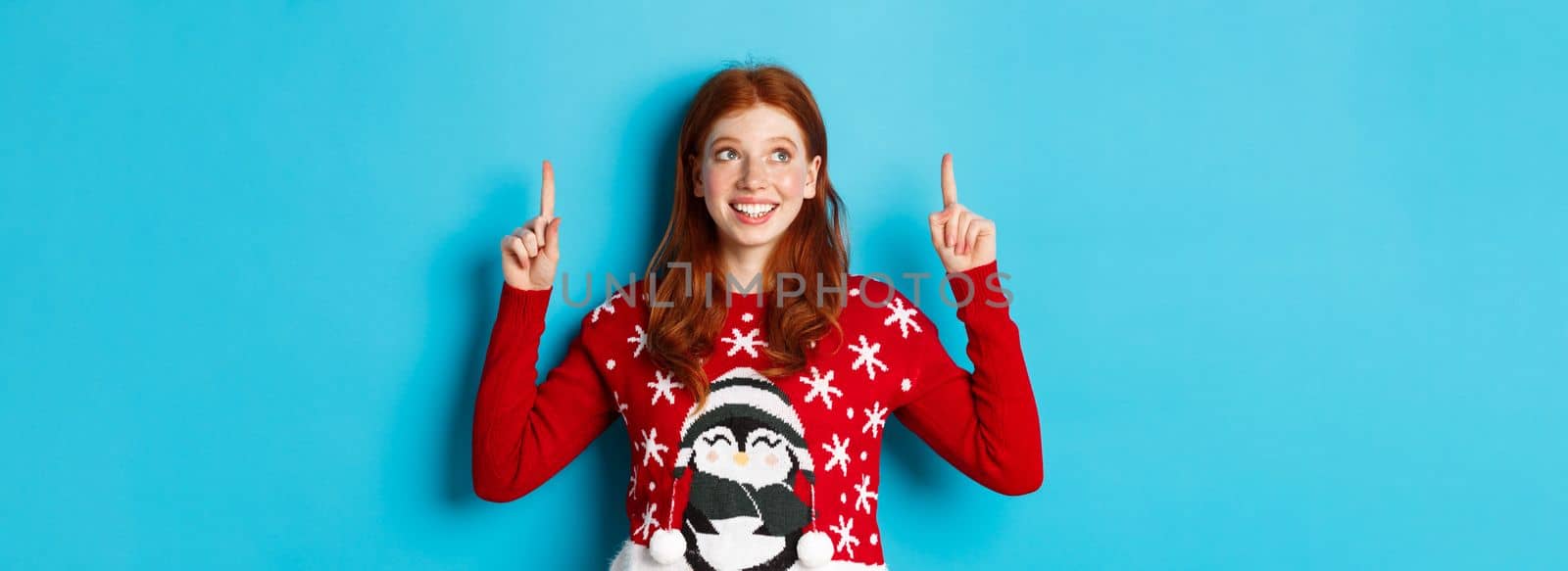 Winter holidays and celebration concept. Cheerful teen girl with red hair, looking dreamy at logo, pointing fingers up, showing advertisement, standing over blue background by Benzoix