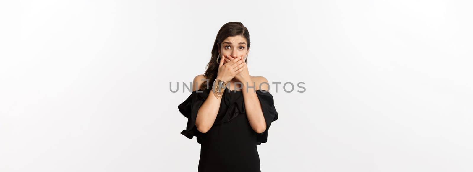 Fashion and beauty. Attractive woman in black dress cover mouth and gasping shocked, staring at camera worried, standing over white background.