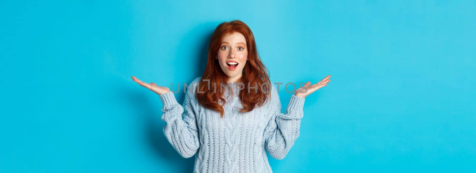 Image of redhead young woman spread hands sidewas, looking with disbelief and amazement, hear news, standing over blue background by Benzoix
