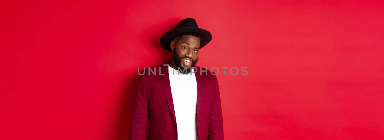 Christmas shopping and people concept. Handsome bearded african american guy looking at camera, smiling confident, wearing party clothes, red background.