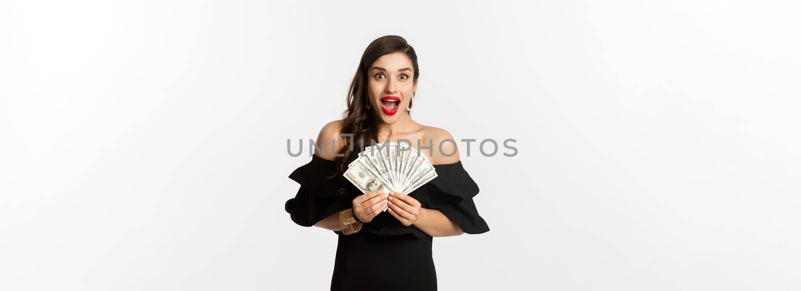 Beauty and shopping concept. Excited woman in black dress, showing money prize and staring happy at camera, standing over white background.