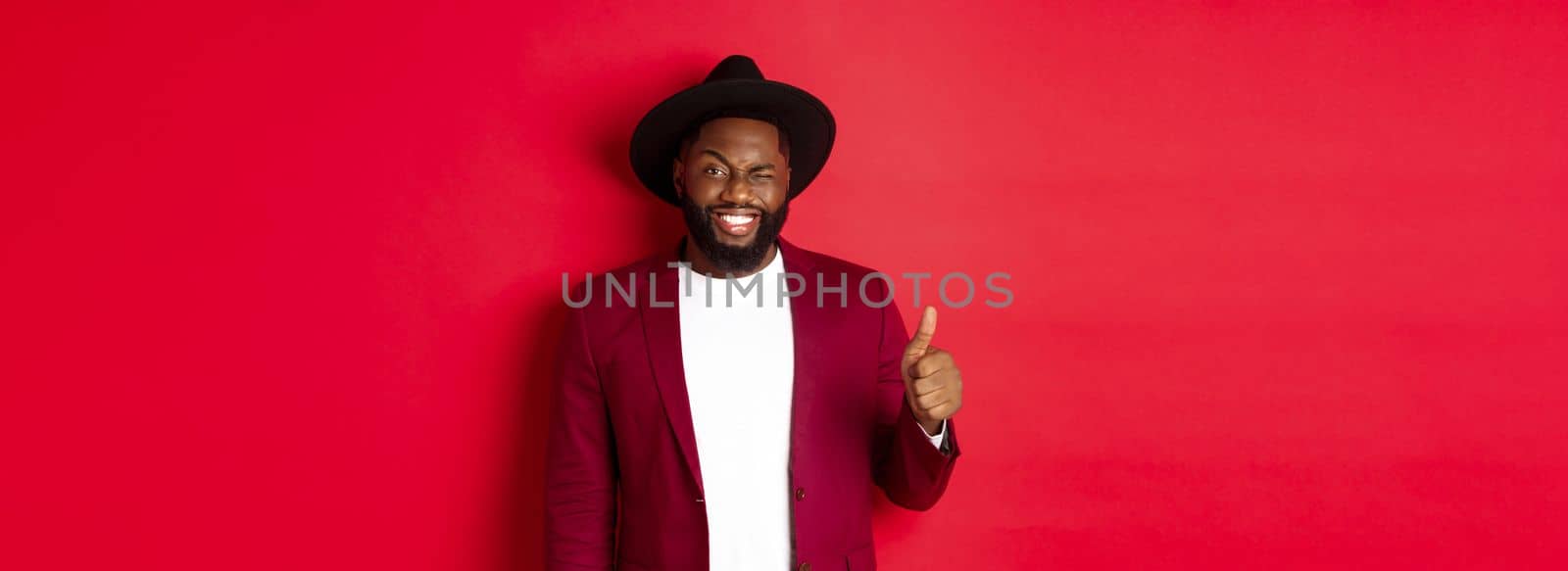 Christmas shopping and people concept. Handsome bearded Black man in party blazer showing thumb up, winking and smiling at camera, recommending promo, red background.