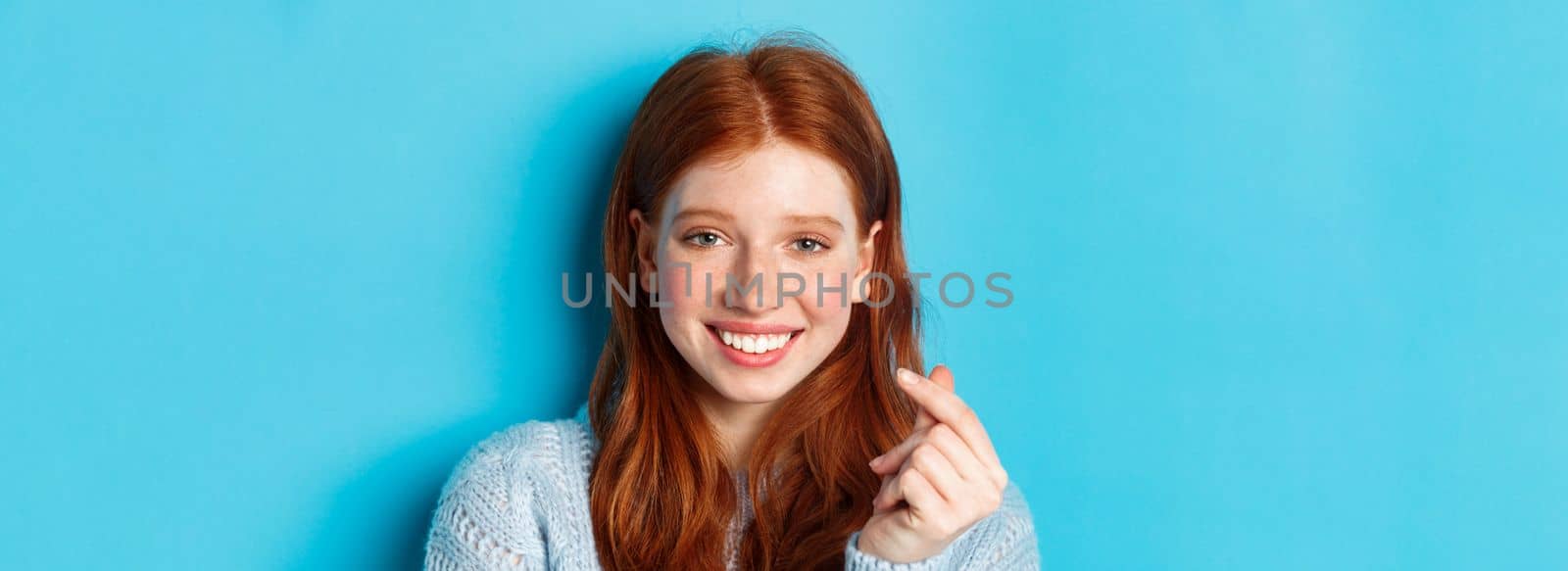 Headshot of cute caucasian woman with red hair and freckles showing heart sign and smiling, standing against blue background.
