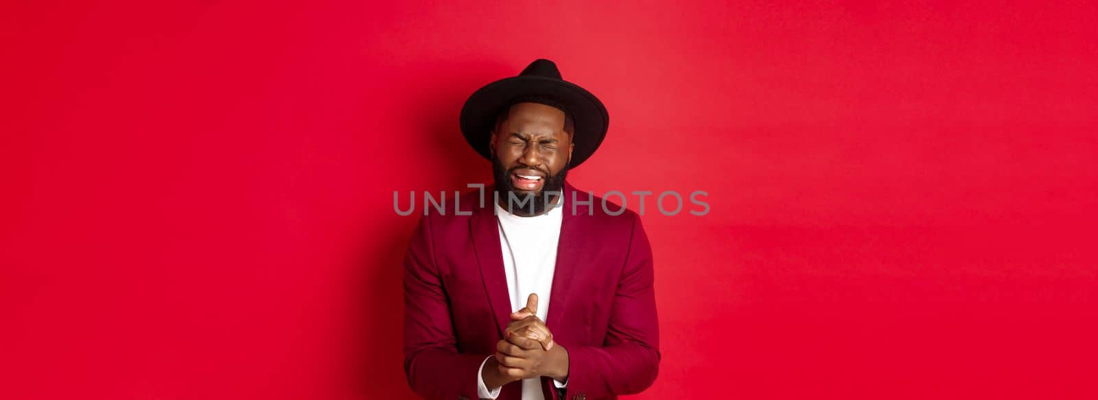 Miserable and heartbroken Black man crying, begging for help, holding hands in pray, standing against red background.