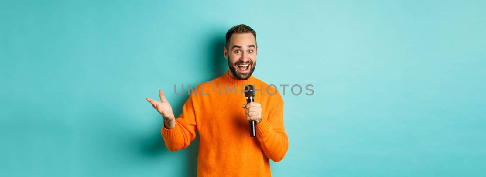 Handsome adult man perform song, singing into microphone, standing against turquoise background.