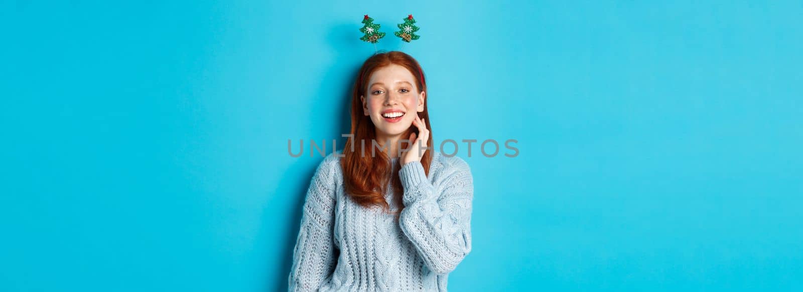 Winter holidays and Christmas sales concept. Beautiful redhead female model celebrating New Year, wearing funny party headband and sweater, smiling at camera by Benzoix