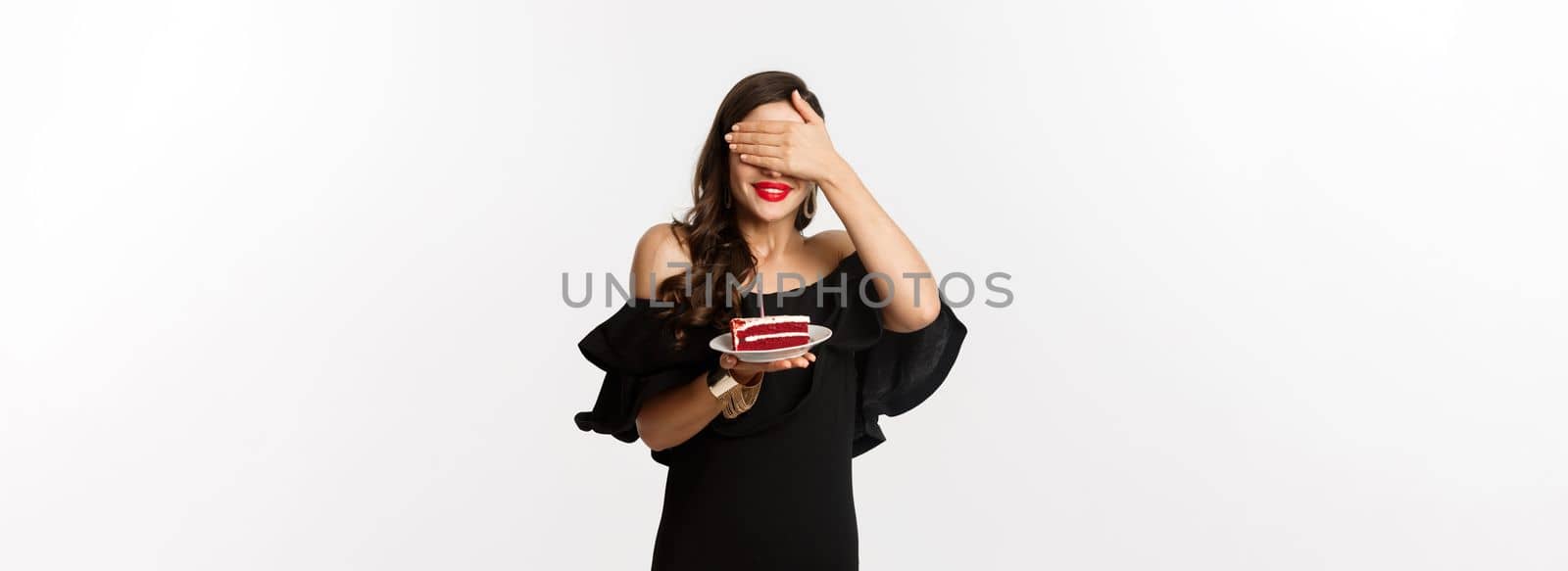 Celebration and party concept. Happy birthday girl in black dress, red lipstick, close eyes and making wish on b-day cake, standing over white background.