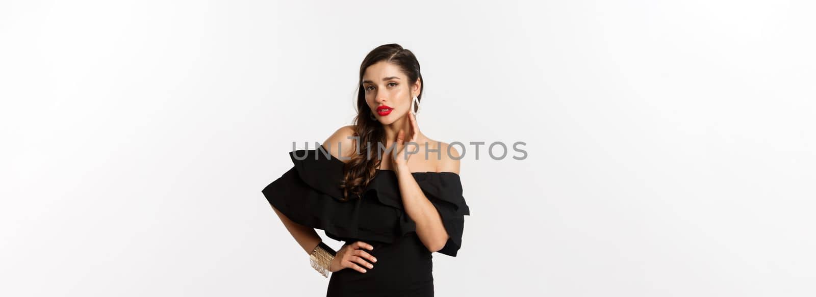 Sensual young woman in black dress, showing her earrings and looking sexy at camera, standing over white background.