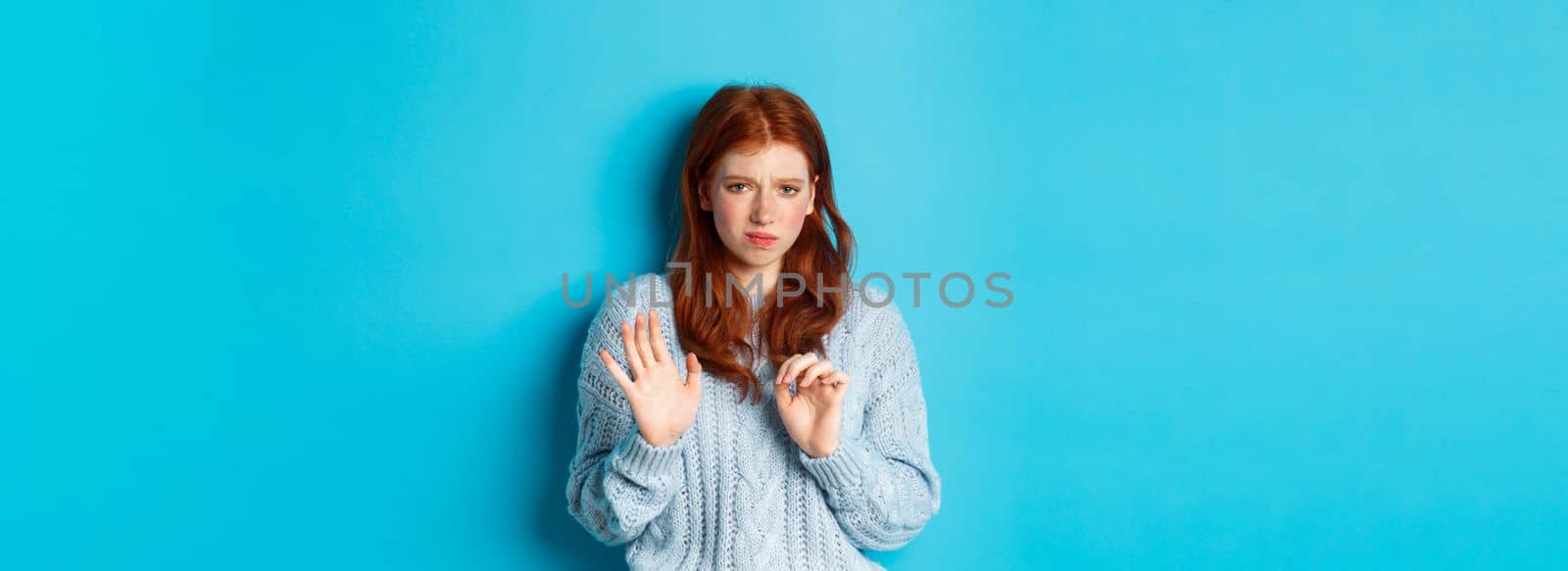 Reluctant redhead girl asking to stay away, shaking hand in rejection gesture, decline offer and grimacing displeased, standing over blue background.