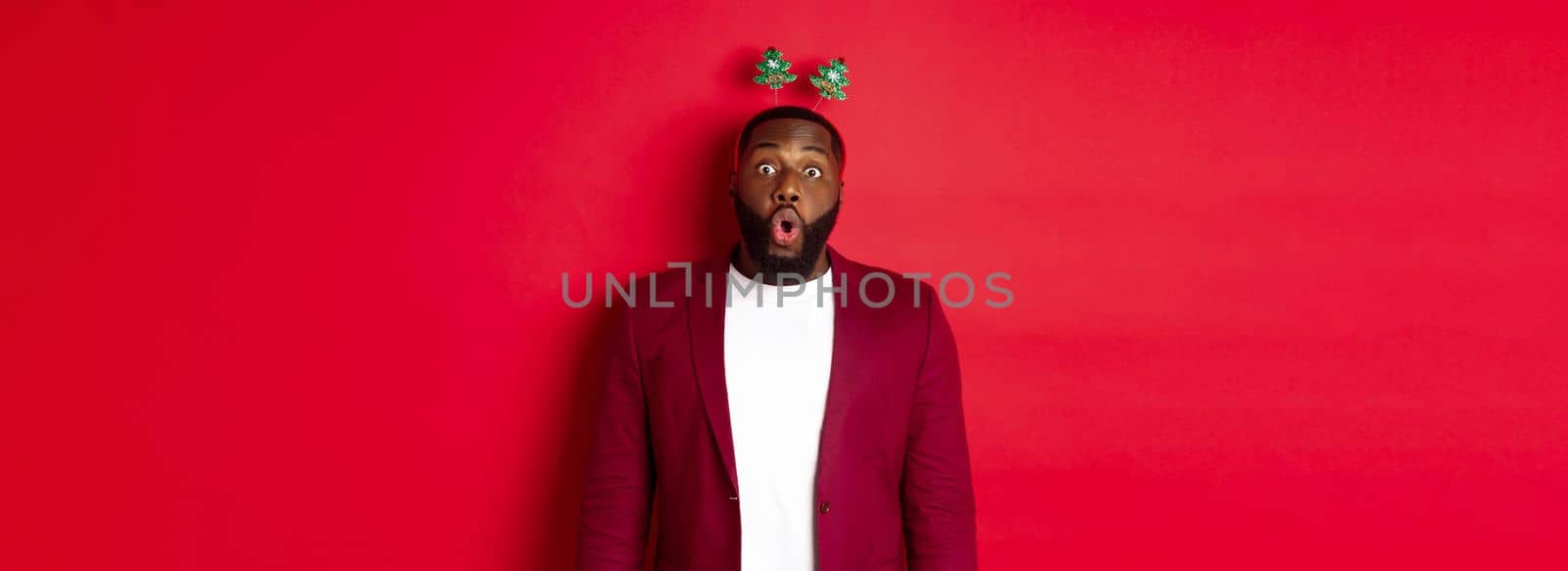 Merry Christmas. Happy african american man celebarting New Year, wearing party headband and making funny faces , standing over red background.