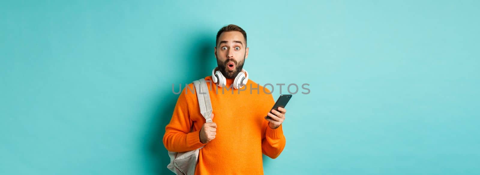 Caucasian man with headphones and backpack staring at camera surprised after reading phone message, standing over turquoise background.