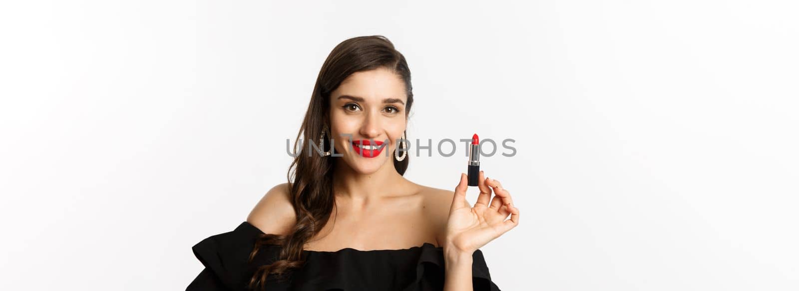 Fashion and beauty concept. Beautiful woman in black dress applying red lipstick and makeup, going on party, standing over white background by Benzoix