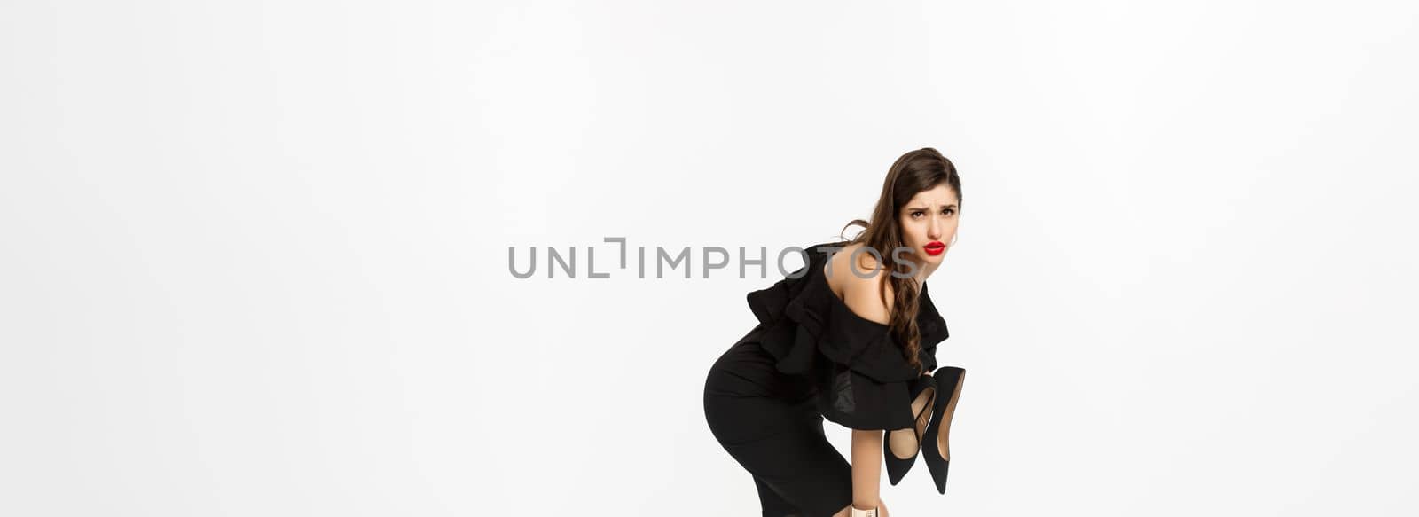 Beauty and fashion concept. Full length of woman feeling pain in feet, take-off high heels and rubbing foot with tired face, standing in black dress over white background.