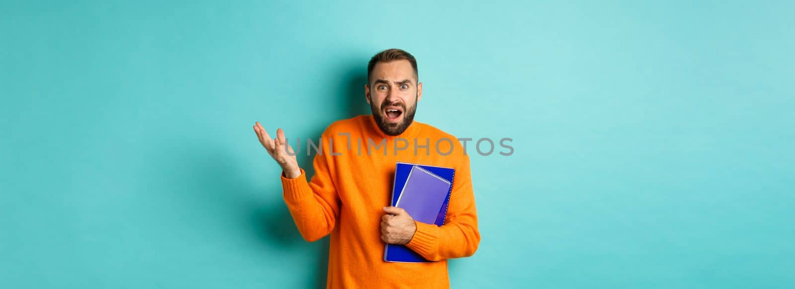 Education. Confused and disappointed man arguing, holding notebooks and complaining, standing over light blue background by Benzoix