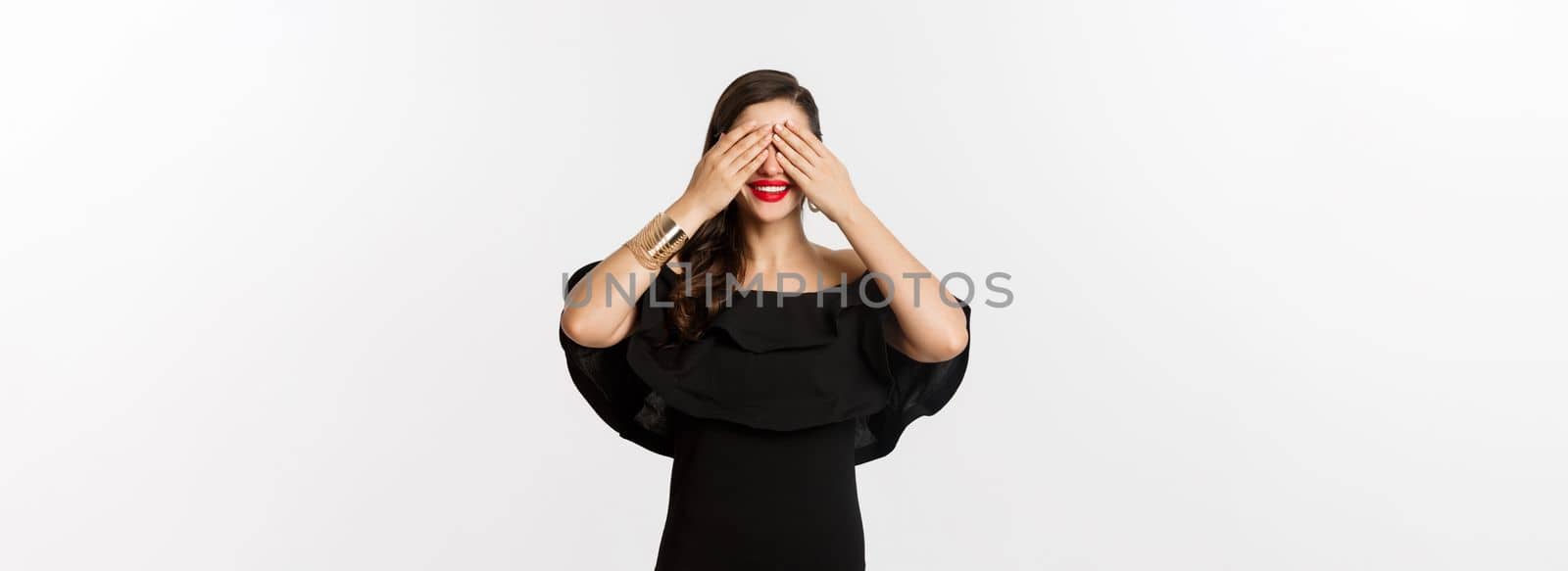 Fashion and beauty. Beautiful woman in black dress and red lipstick, covering eyes and smiling, waiting for surprise, standing over white background by Benzoix