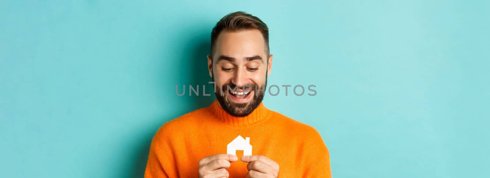 Real estate concept. Happy young man searching for home rent, holding house paper maket and smiling, standing over blue background by Benzoix