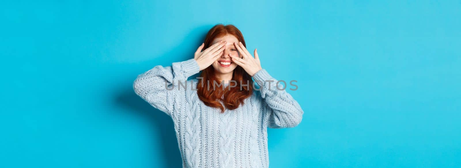 Excited teenage redhead girl open eyes to see holiday surprise, receiving presents, looking amazed at camera, standing over blue background.