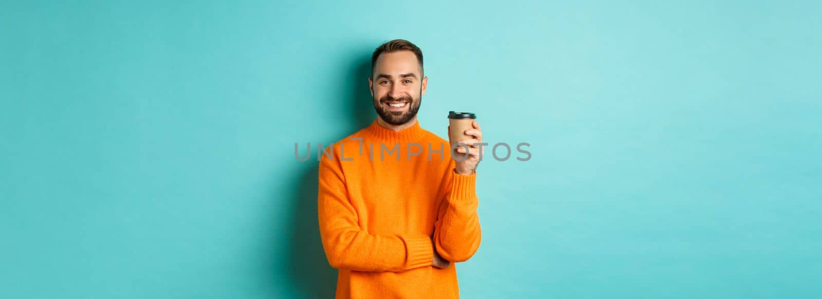 Handsome smiling man taking break, drinking coffee from takeaway, standing over blue background.