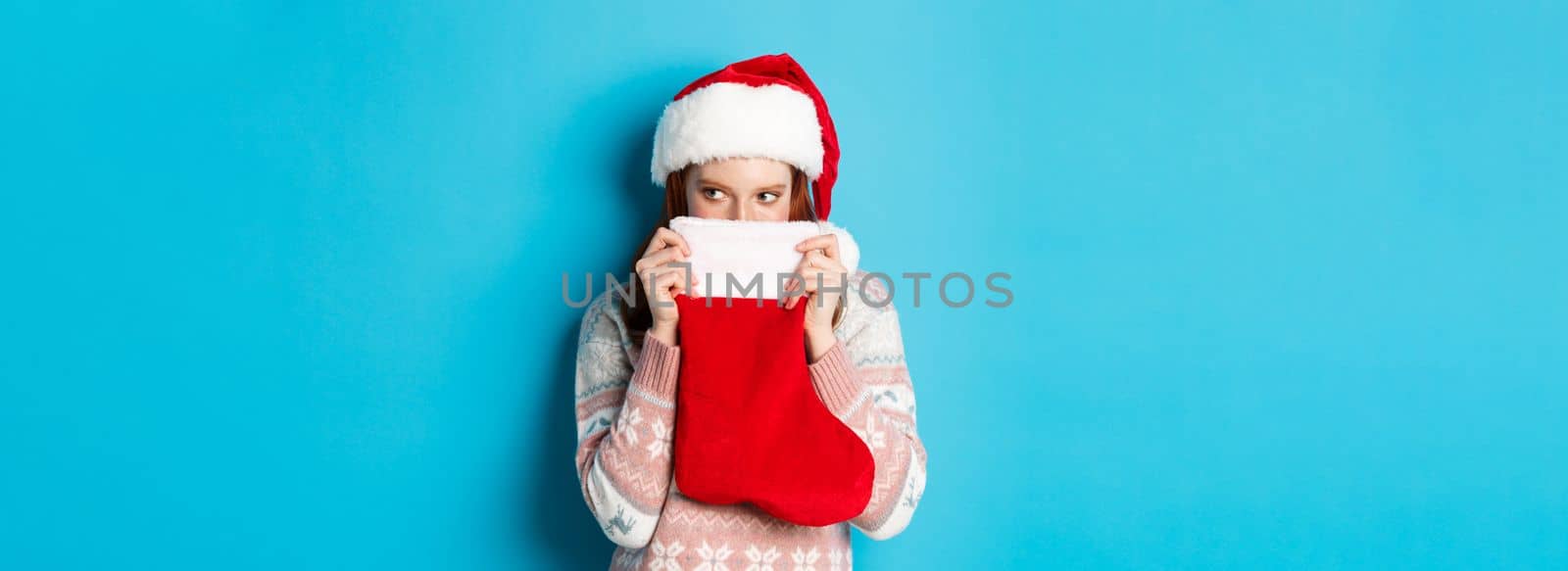 Cute girl cover face with christmas stocking, staring right with cunning gaze, standing in Santa hat and celebrating winter holidays, blue background.