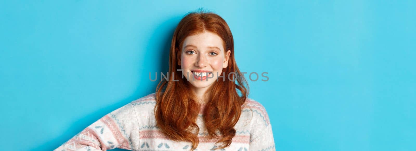 Close-up of attractive female student with red hair, wearing winter sweater, smiling happy and staring at camera, blue background.
