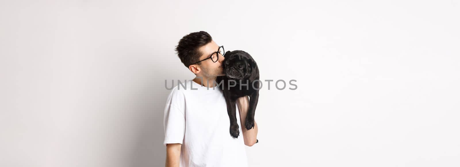 Image of handsome young man kissing his cute black pug, holding dog on shoulder, standing over white background.