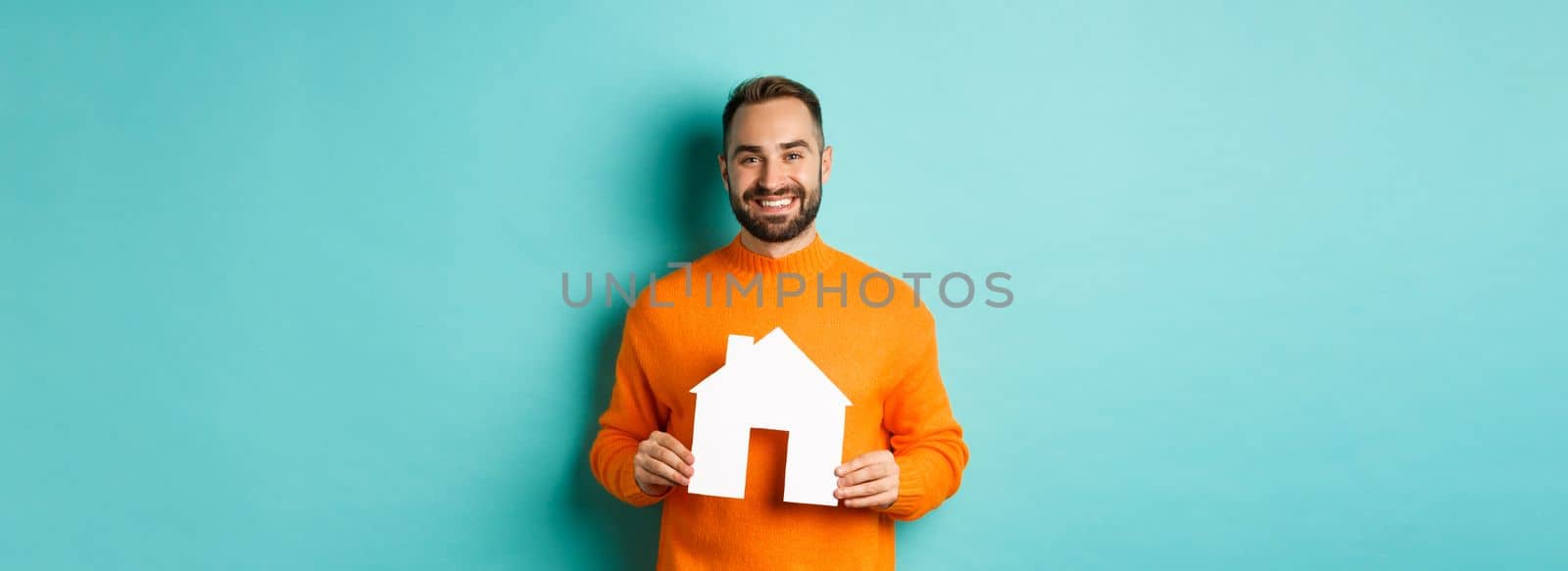 Real estate concept. Happy young man searching for home rent, holding house paper maket and smiling, standing over blue background.