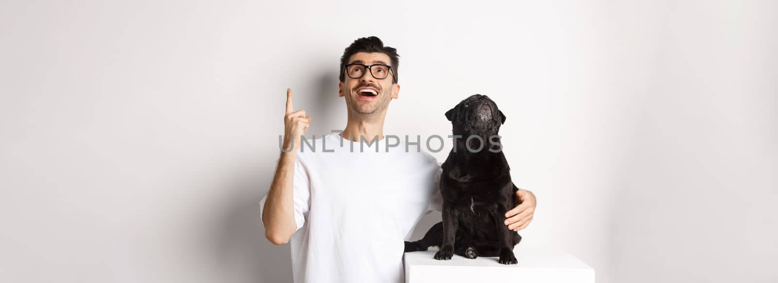 Image of happy young man and dog both looking up at promo, owner pointing finger at top, pug staring at loog, standing over white background by Benzoix