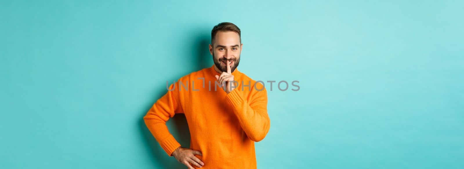 Happy man asking keep voice down, shushing and smiling, prepare surprise, hushing at camera, standing over turquoise background.