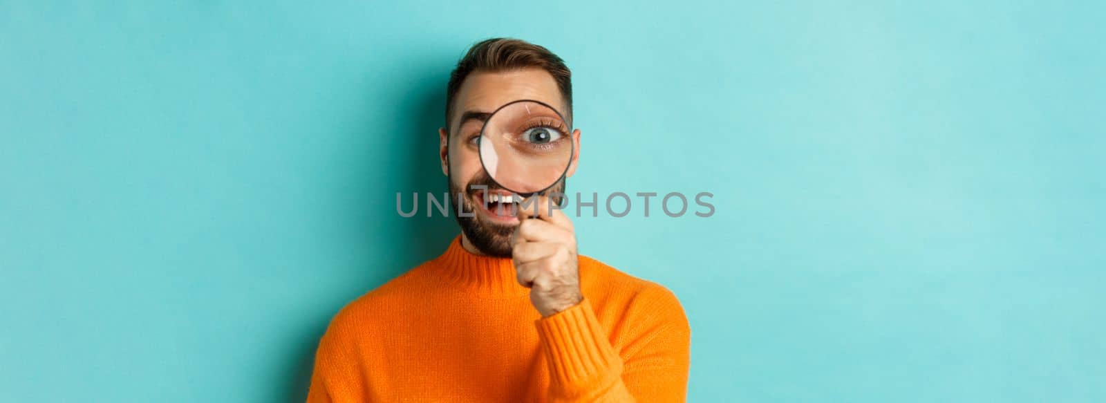 Funny man looking through magnifying glass, searching or investigating something, standing in orange sweater against turquoise background.