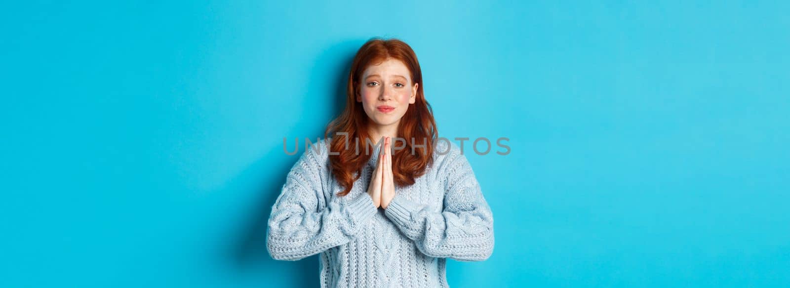Cute teenage redhead girl asking for help, smiling while begging for favour, need something, standing over blue background.