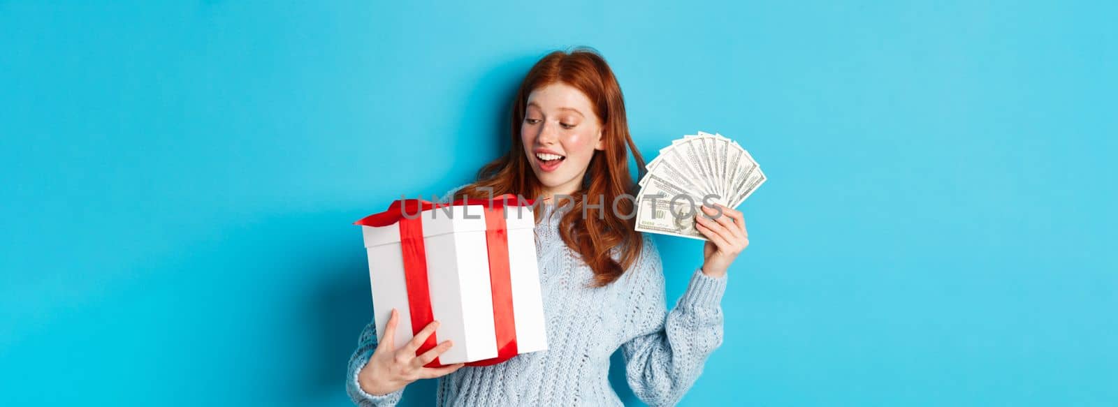 Christmas and shopping concept. Cheerful girl with red hair, holding money and big New Year gift, smiling happy, standing over blue background.