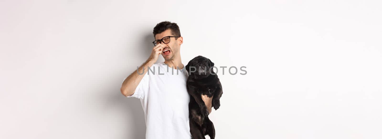 Dog owner squinting and shutting nose from disgusting reek, holding pug with bed smell, standing over white background.