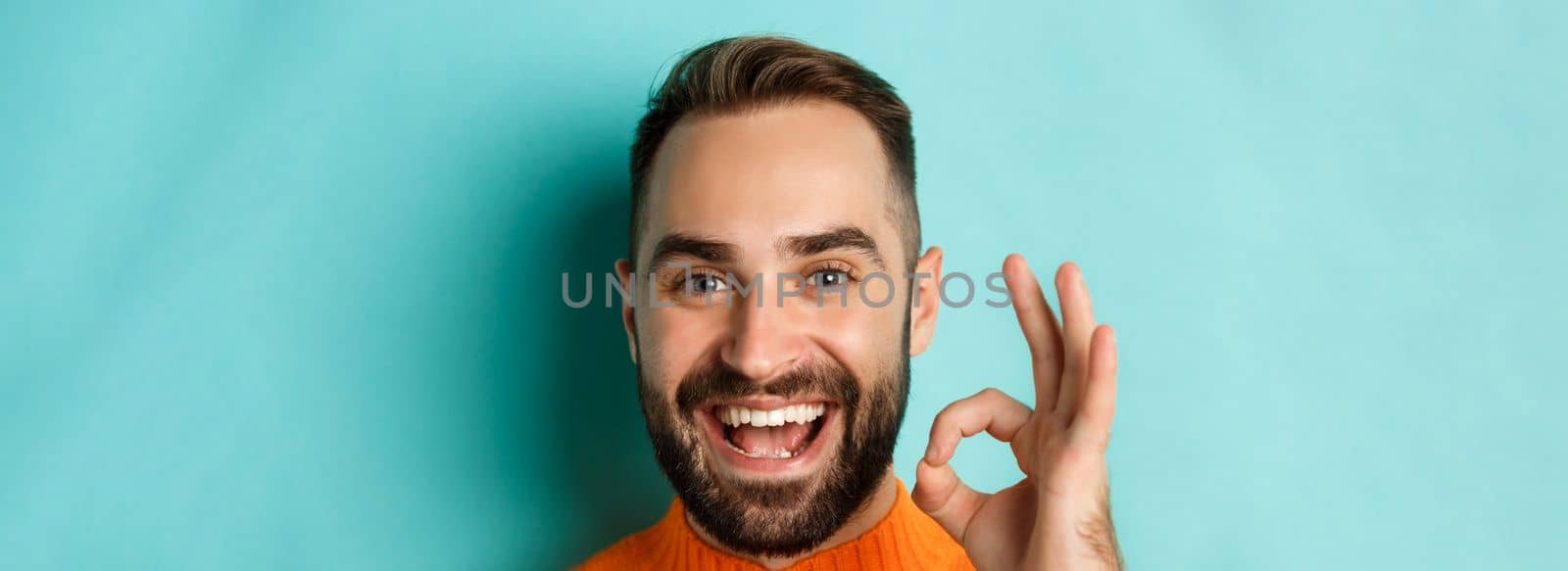 Close-up of confident and happy man with beard showing okay sign, approve and like, giving positive reply or agree, standing over turquoise background.