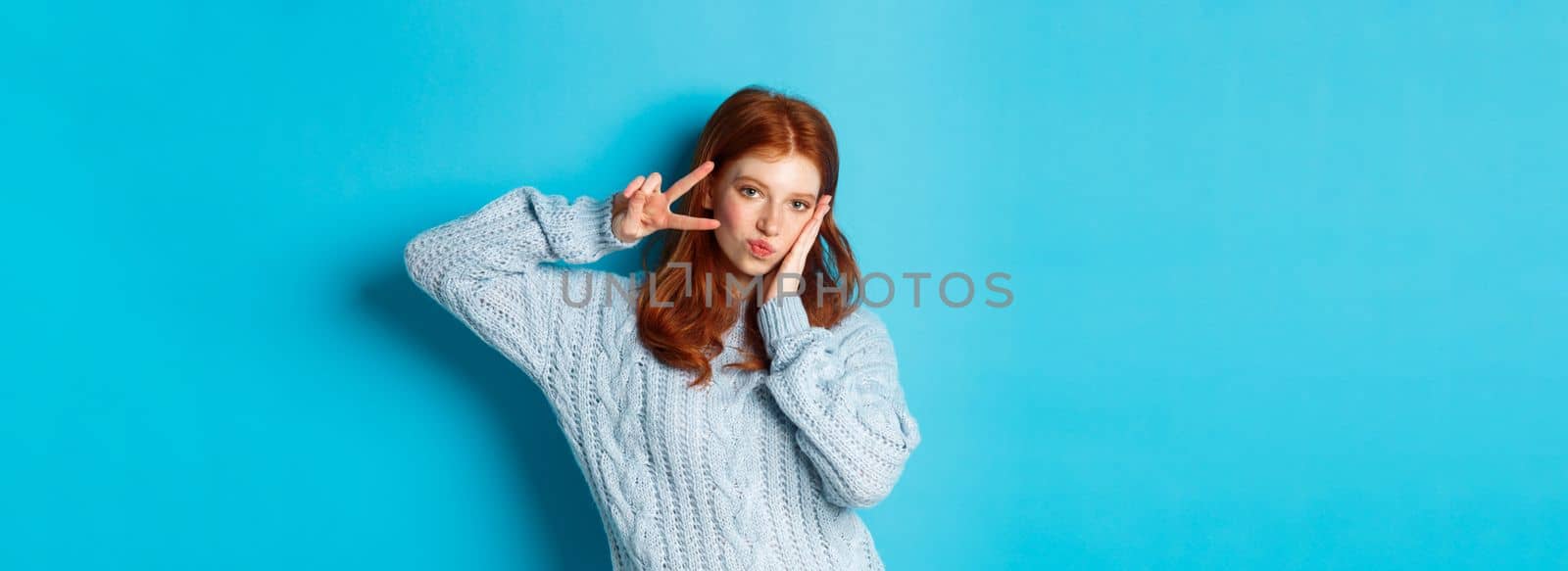 Modern teen girl with red hair, showing peace sign and posing in sweater against blue background.