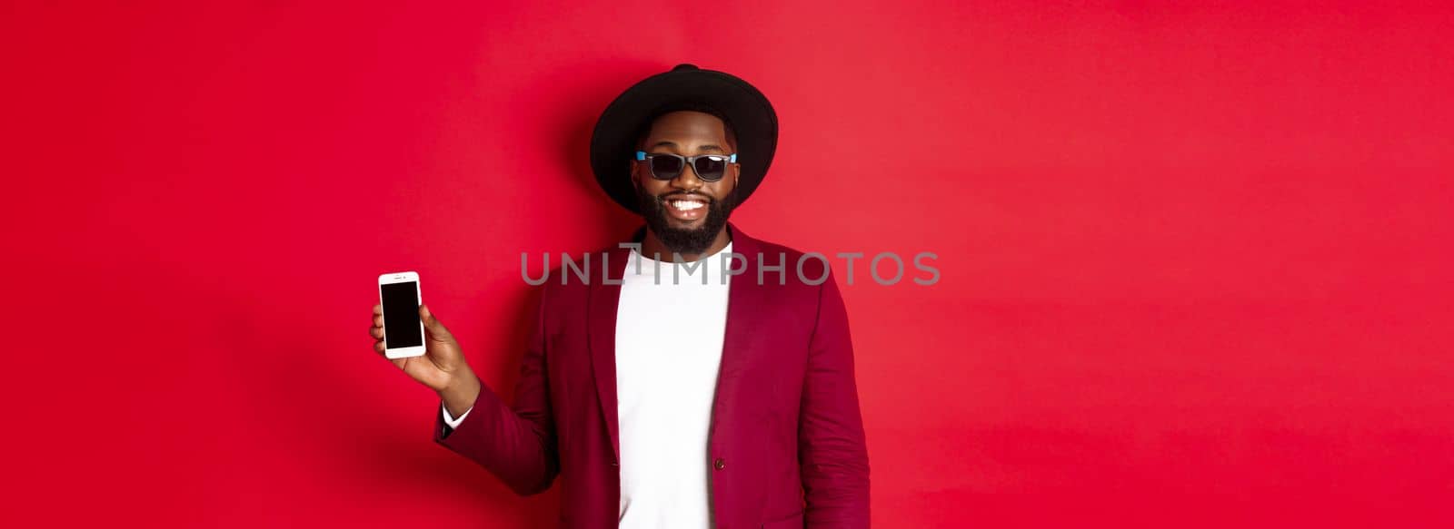 Handsome and stylish Black man showing phone screen at camera, recommending online store or application, standing over red background.