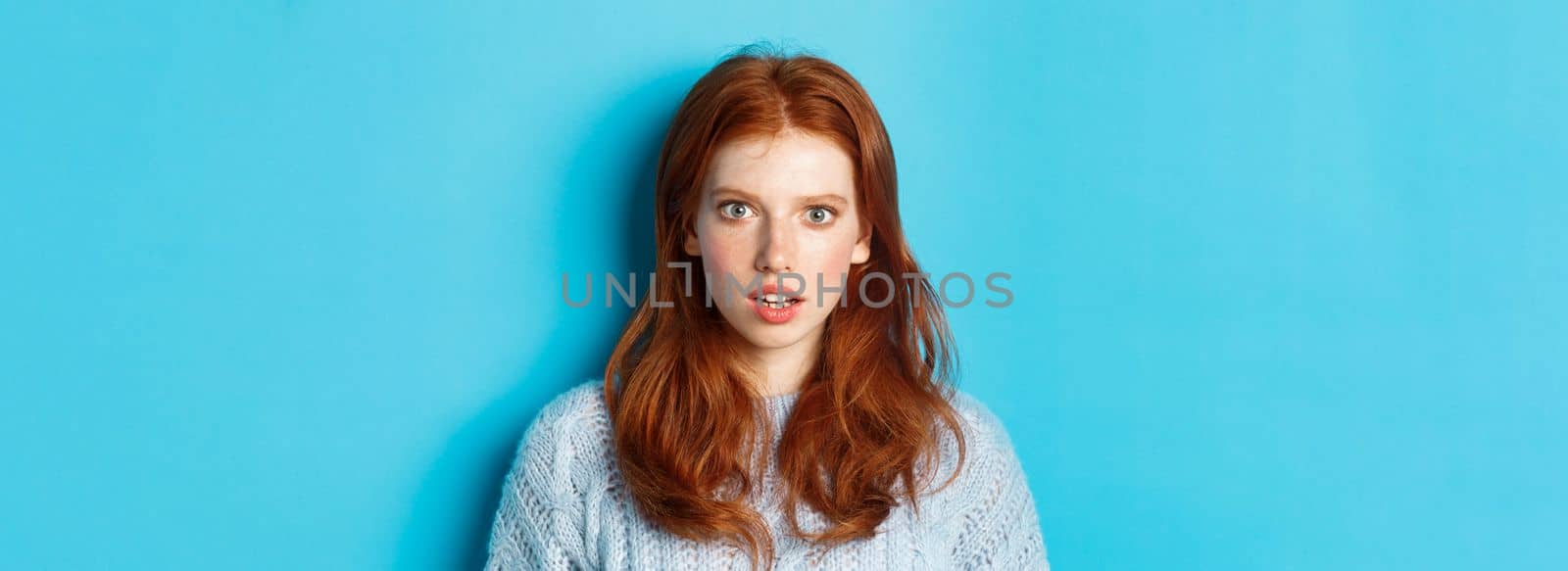 Close-up of amazed redhead girl staring at camera with complete disbelief, standing shocked against blue background by Benzoix