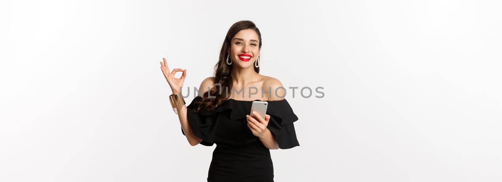 Online shopping concept. Attractive woman in trendy black dress, makeup, showing okay sign in approval and using mobile phone app, white background.