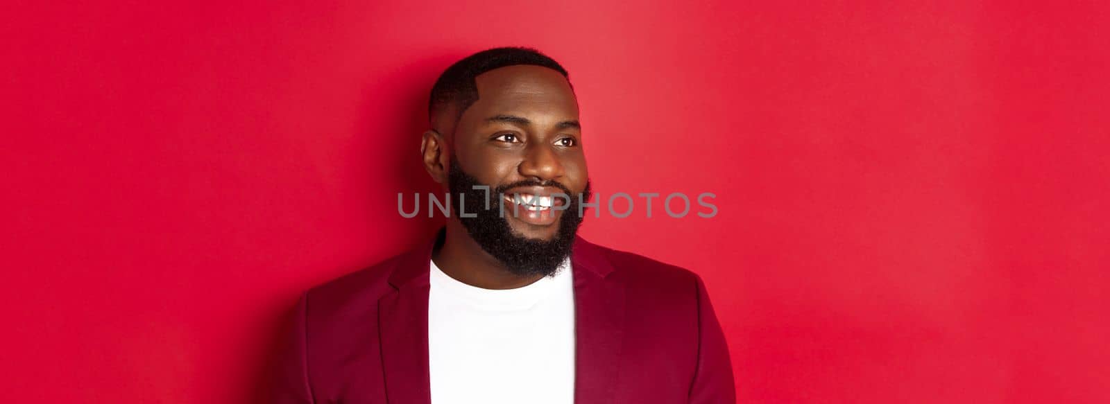 Close-up of handsome african american man with beard, looking left and smiling joyful, standing over red background.