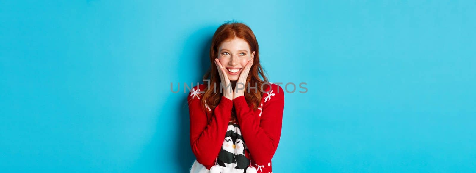 Winter holidays and Christmas Eve concept. Adorable redhead girl blushing and touching cheeks from happiness, smiling and looking at upper left corner promo, blue background.