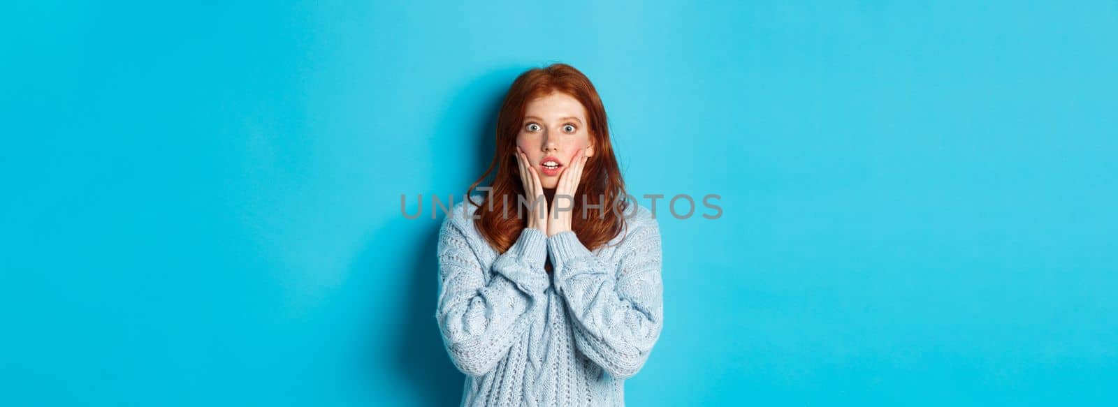 Shocked redhead girl staring at camera speechless, express disbelief and amazement, standing in sweater against blue background.