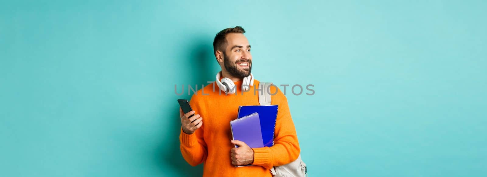 Education. Handsome male student with headphones and backpack, using mobile phone and holding notebooks, smiling happy, standing over turquoise background.