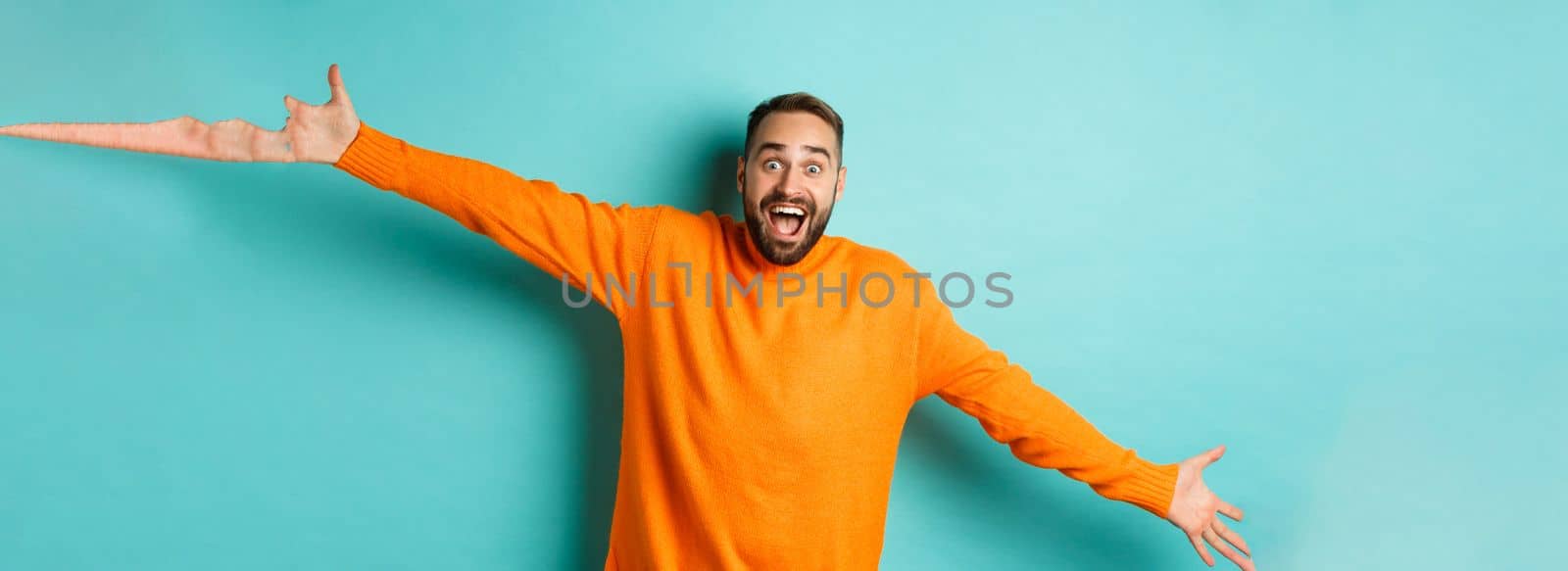 Surprised and happy man spread hands in warm welcome, reaching for hug, greeting you, showing something big, standing over light blue background.