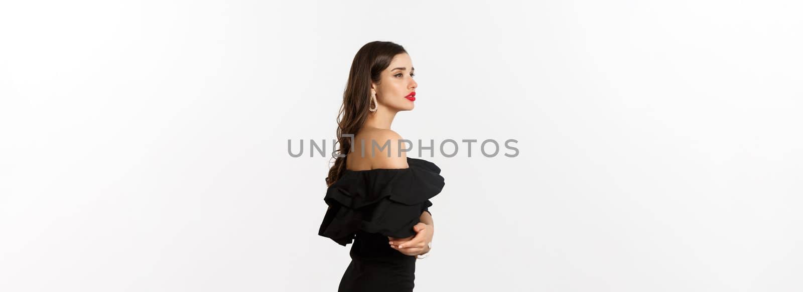 Profile view of elegant young woman with red lips, makeup and black dress, looking dreamy in distance, standing over white background.