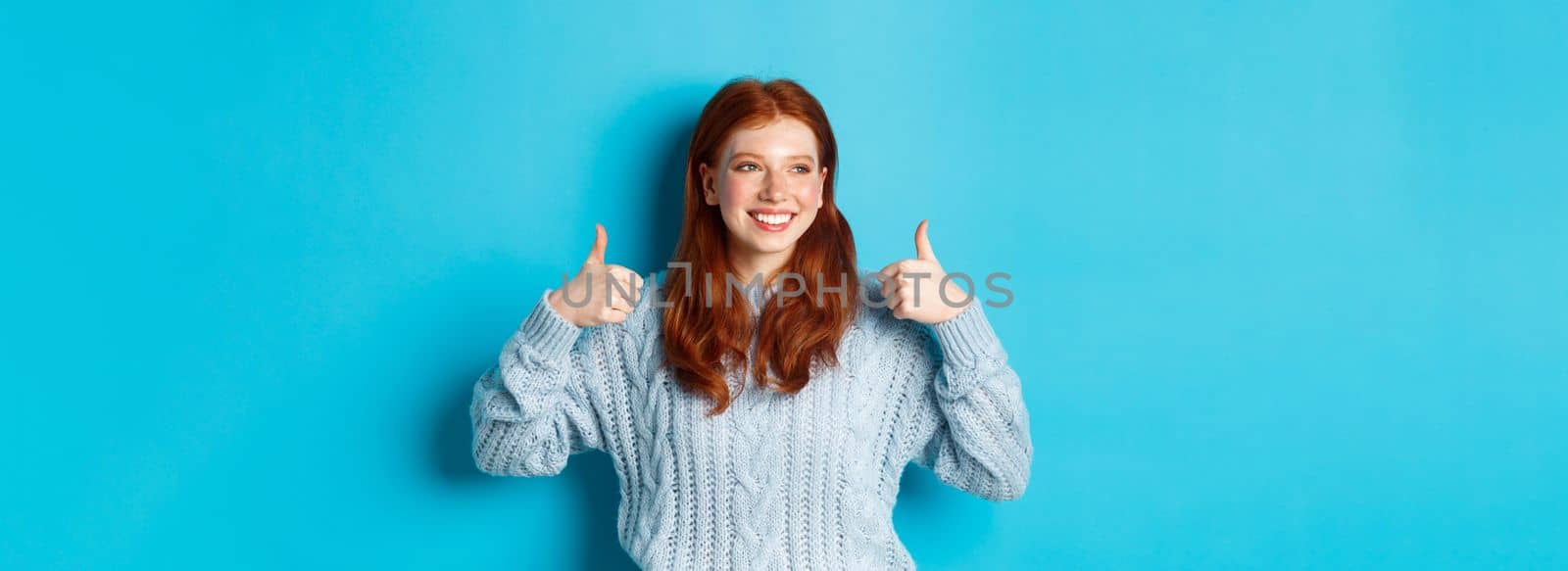Happy smiling woman with red hair, showing thumbs up and looking left satisfied, praising good choice, agree and approve, standing over blue background by Benzoix