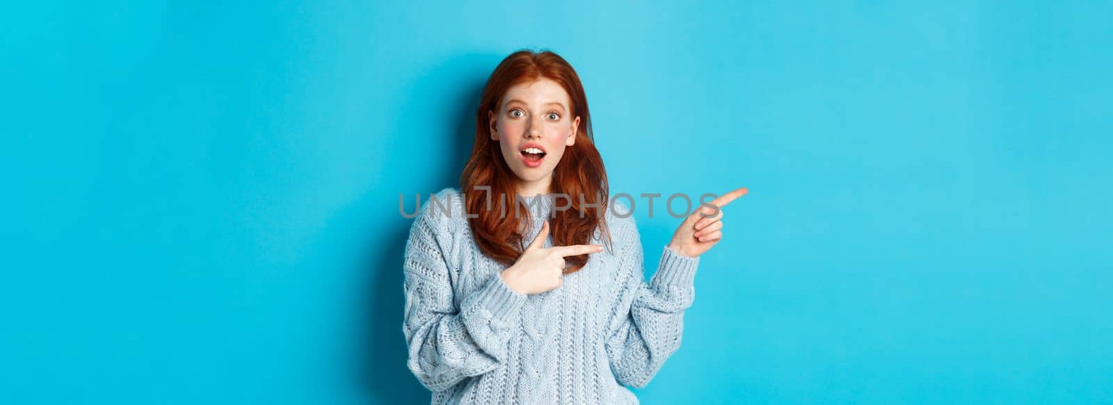Amazed redhead girl in sweater, pointing fingers left at logo and staring excited at camera, standing against blue background.